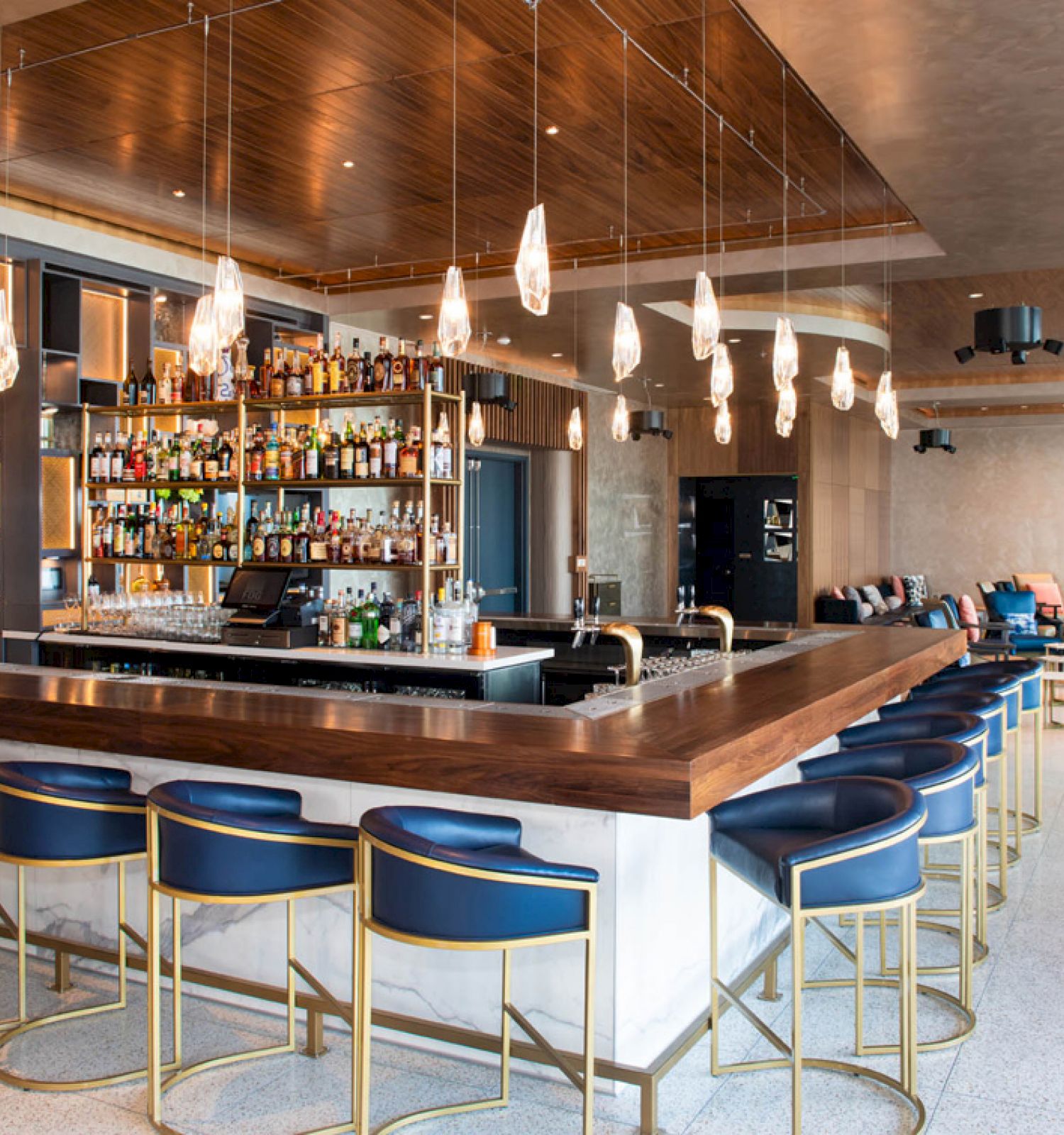 The image shows a modern bar with blue stools, hanging lights, and a well-stocked selection of bottles behind the counter, offering a cozy setting.
