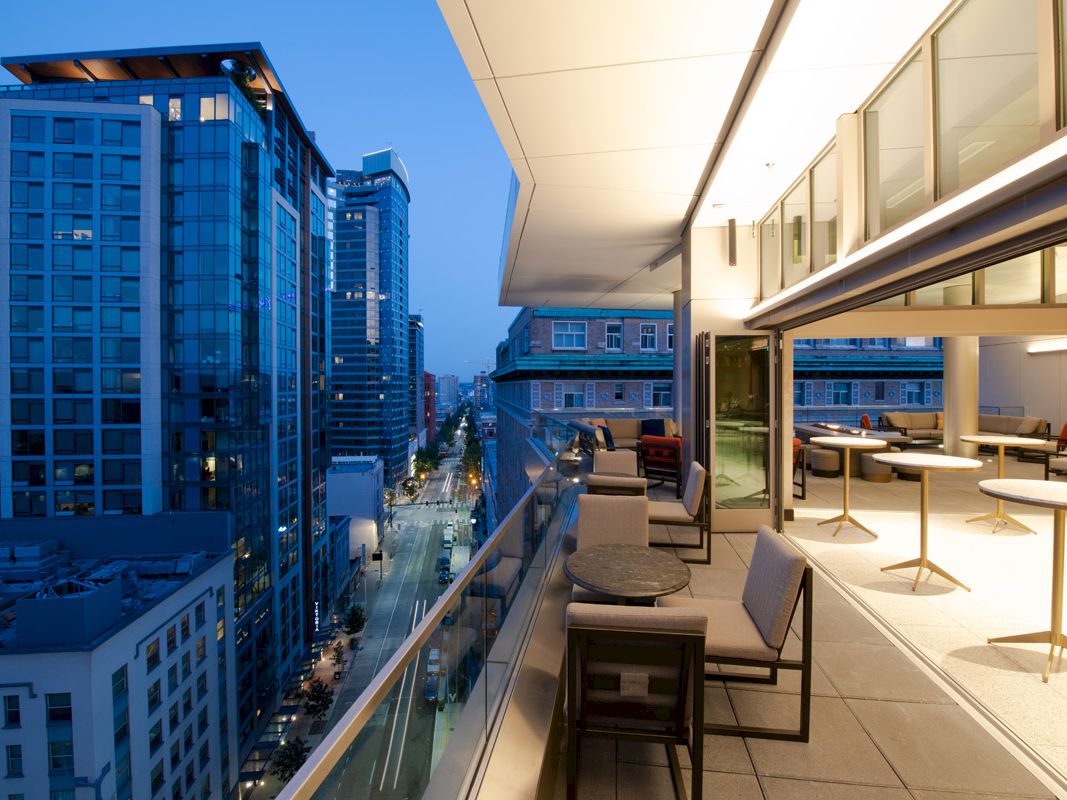 A modern rooftop terrace with seating overlooks a cityscape at dusk, featuring tall buildings and a street with lights on.