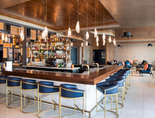 A modern bar with a wooden countertop, blue stools, hanging lights, and a wide selection of spirits. Seating areas are visible in the background.