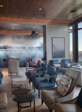 The image shows a modern lounge with many chairs, large windows offering a scenic view of water and a skyline mural on the wall.