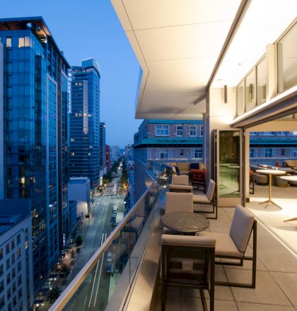 A modern cityscape at dusk with a rooftop terrace featuring seating, tables, and a view of surrounding buildings and a street below.