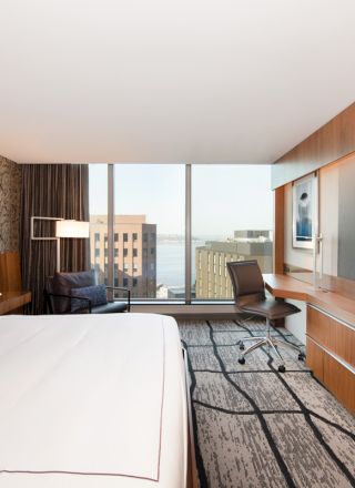 A modern hotel room with a large bed, desk, chair, TV, and view through a window overlooking city buildings and a body of water.