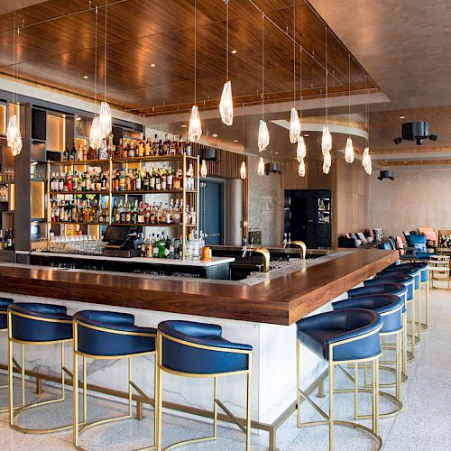 The image shows a modern bar with a stocked liquor shelf, blue bar stools, hanging lights, and a seating area in the background.