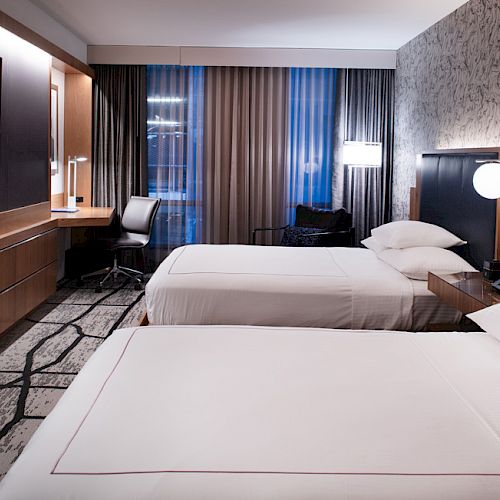 The image shows a modern hotel room with two beds, a desk, a chair, a television, and stylish decor with ambient lighting on the walls.