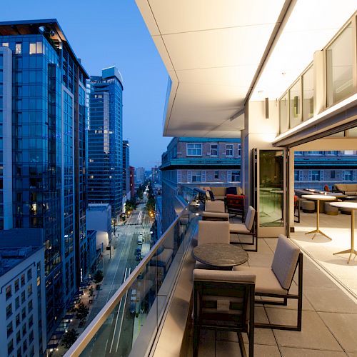 A modern urban balcony with seating overlooks a city street, flanked by tall buildings in the evening light.
