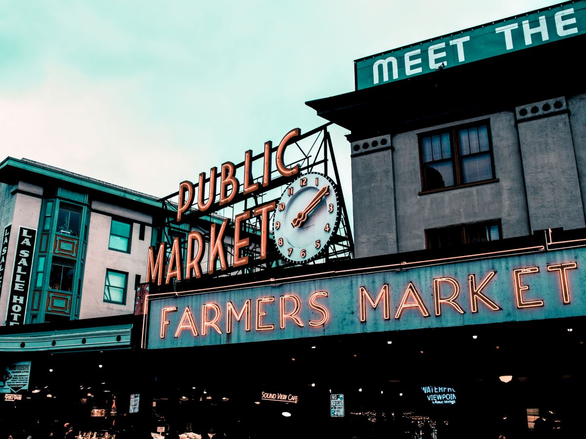 The image shows a public market sign with a clock and neon letters reading "Farmers Market," set against the background of urban buildings.
