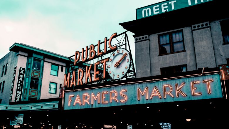 The image shows a public market sign with a clock and neon letters reading "Farmers Market," set against the background of urban buildings.