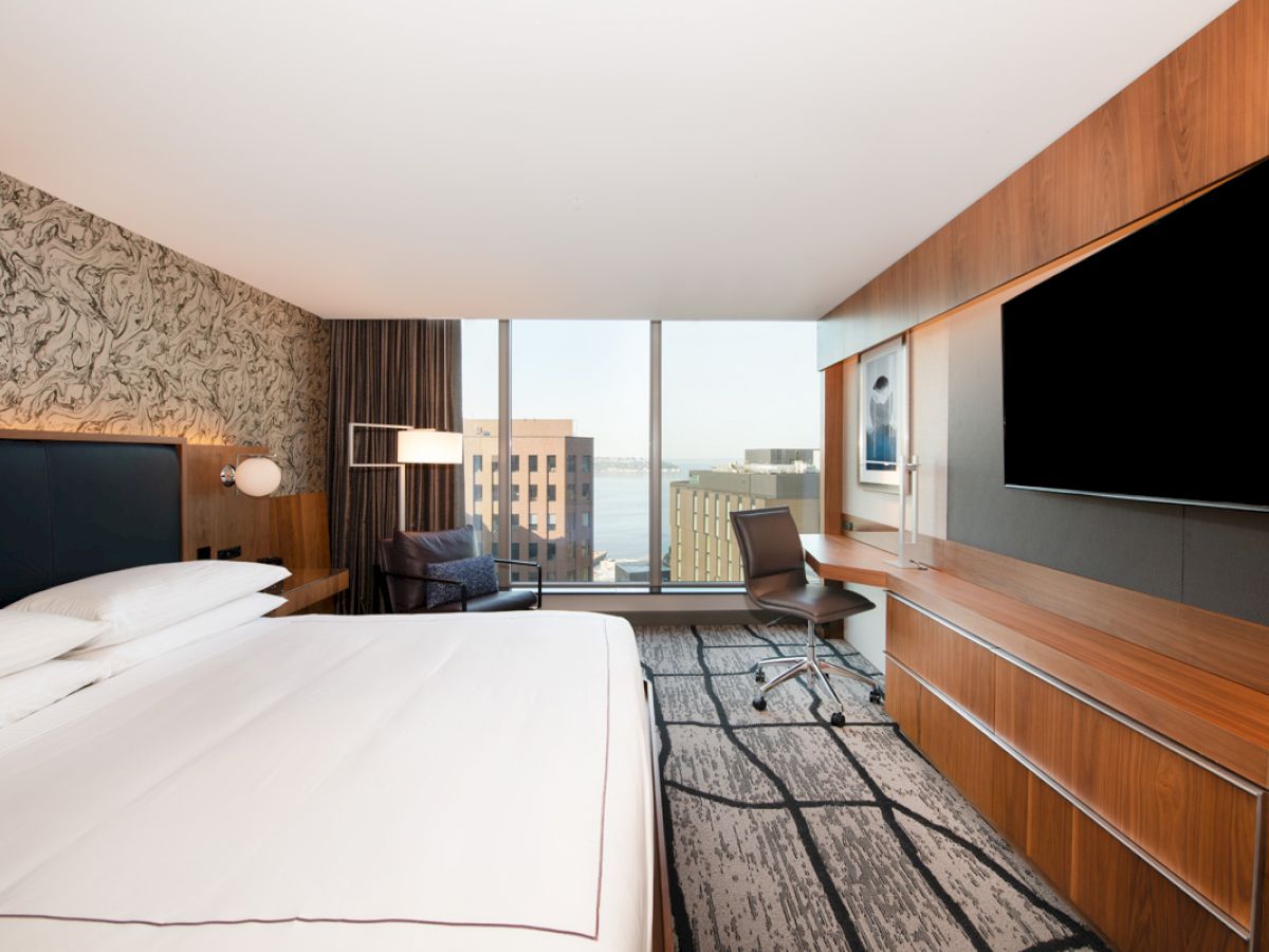 A modern hotel room with a large bed, desk, TV, and a window view of city buildings and water in the background.