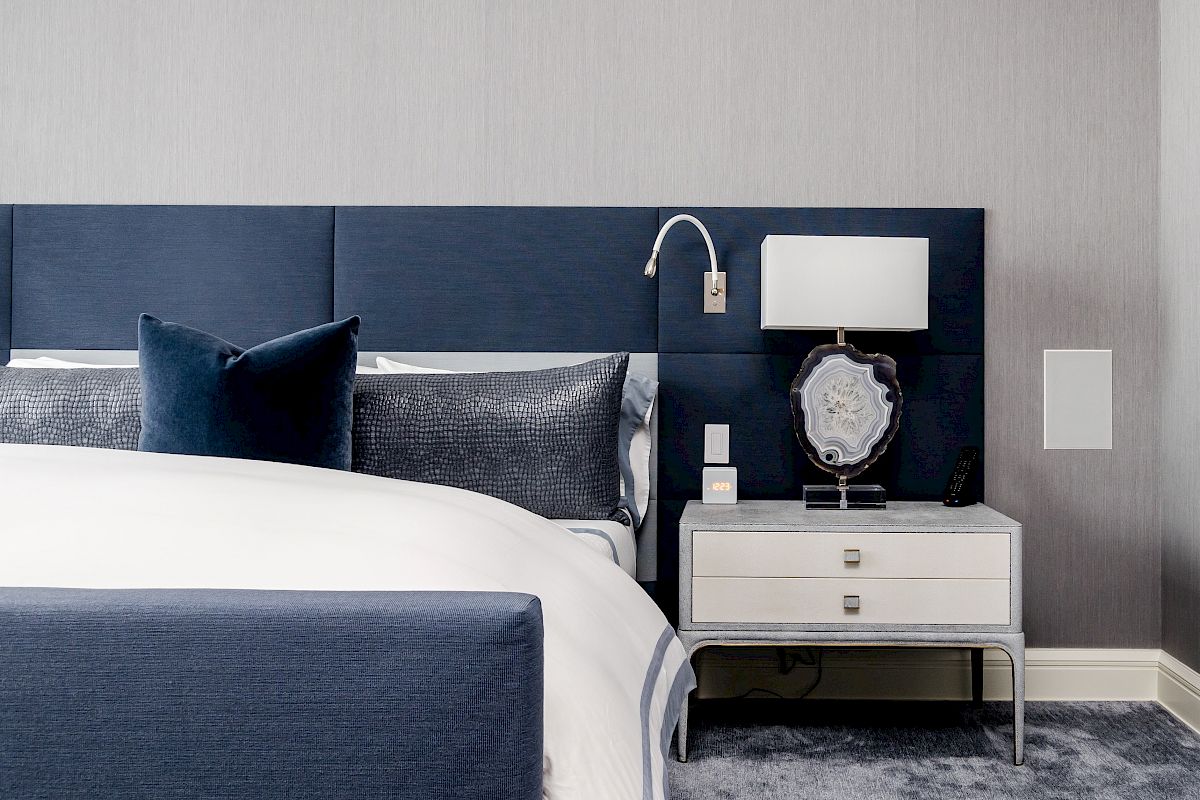 A modern bedroom with a blue upholstered bed, decorative pillows, and a sleek nightstand with a lamp and decor piece.