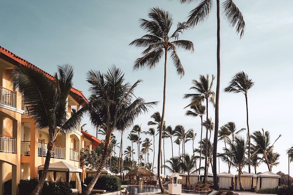 A tropical resort pool area with lounge chairs, palm trees, and adjacent buildings under a clear sky.