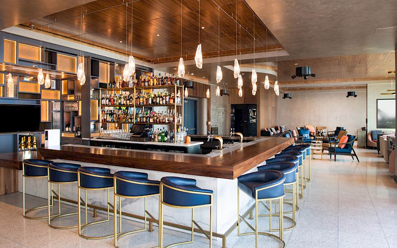 A stylish bar with a wooden counter, blue stools, hanging lights, and a display of bottles. Modern lounge seating in the background.