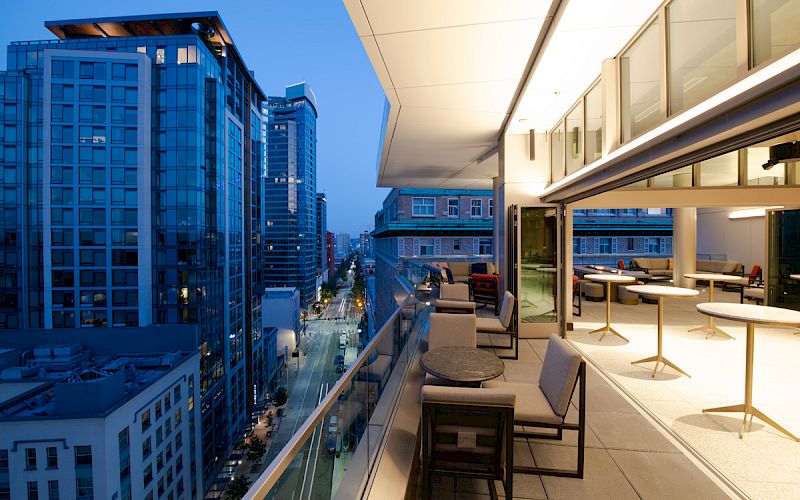 A modern rooftop patio with seating overlooks a cityscape at dusk, featuring high-rise buildings and street views.