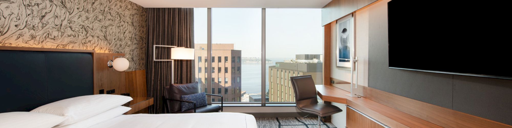 The image shows a modern hotel room with a large bed, a flat-screen TV, a desk, and a city view through a large window.